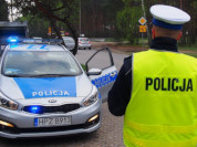 Na zdjęciu po prawej stronie znajduje się oznakowany radiowóz z włączonymi błyskami, po lewej stojący tyłem umundurowany policjant ruchu drogowego.