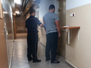 Na zdjęciu widać umundurowanego policjanta, trzyma mężczyznę przed drzwiami do policyjnej celi.
