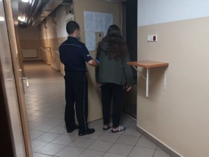 Na zdjęciu widać umundurowanego policjanta, który trzyma otwarte do policyjnej celi metalowe drzwi. Przed policjantem znajduje się odwrócona tyłem kobieta.