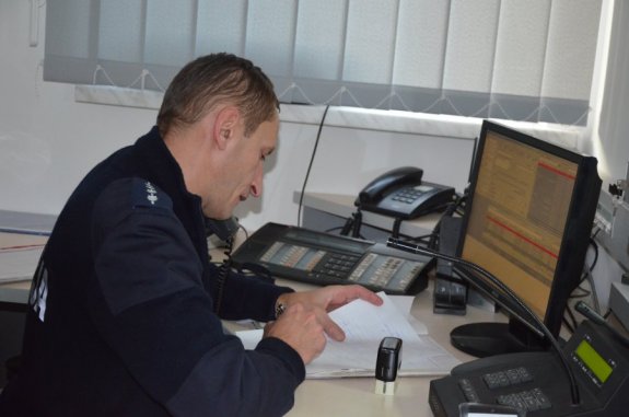 Na zdjęciu widać umundurowanego policjanta, który siedzi przy biurku, na którym znajduje się monitor komputera i telefony stacjonarne.