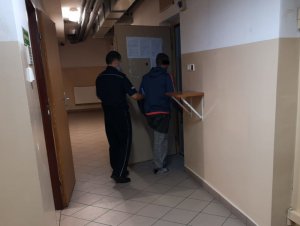 Na zdjęciu widać umundurowanego policjanta, który trzyma otwarte metalowe drzwi celi. Przed nim jest mężczyzna, który wchodzi do środka pomieszczenia.