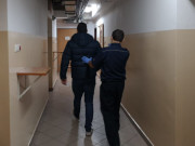 Na zdjęciu widać umundurowanego policjanta, który prowadzi przed sobą wzdłuż korytarza mężczyznę. Policjant na dłoniach ma niebieskie lateksowe rękawiczki.