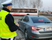 Na zdjęciu, po prawej stronie jest umundurowany policjant drogówki, który stoi przy nieoznakowanym radiowozie. W tle widać autobus i drogę.