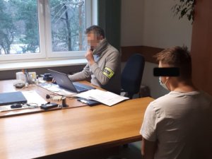 Na zdjęciu widać nieumundurowanego policjanta, który siedzi przy biurku. Na biurku jest laptop i dokumenty. Z drugiej strony biurka siedzi odwrócony tyłem mężczyzna.