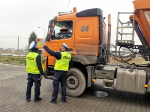 Na zdjęciu widać dwóch umundurowanych policjantów, którzy przeprowadzają kontrolę drogową kierującego samochodem ciężarowym koloru pomarańczowego.