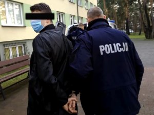 Na zdjęciu widać po lewej stronie mężczyznę z kajdankami założonymi na ręce trzymane z tyłu oraz po prawej umundurowanego policjanta, który prowadzi mężczyznę. Obaj są odwróceni tyłem.