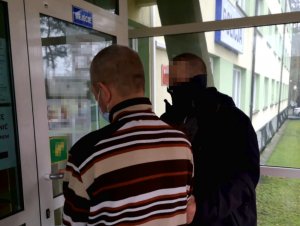 Na zdjęciu widać nieumundurowanego policjanta, który trzyma mężczyznę. Mężczyźni znajdują się przed wejściem do budynku komendy.