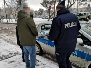 Na zdjęciu widać umundurowanego policjanta i mężczyznę z kajdankami założonymi na ręce trzymane z przodu. Obaj stoją przy oznakowanym radiowozie.