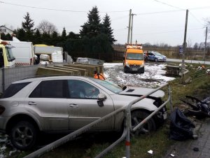 Na zdjęciu widać pojazd koloru szarego, który ma rozbity przód. W tle widać oznakowany radiowóz oraz pojazd pomocy drogowej.