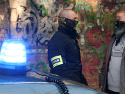 Na zdjęciu widać nieumundurowanego policjanta, przed który stoi zwrócony do niego przodem mężczyzna. Obaj stoją za nieoznakowanym radiowozem.