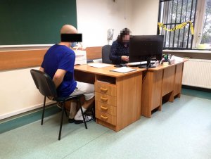 Na zdjęciu widać siedzącego przy biurku mężczyznę, który ma założone kajdanki na ręce trzymane z tyłu. Po drugiej stronie biurka, naprzeciwko mężczyzny, siedzi policjantka. Na biurku jest komputer i są dokumenty.