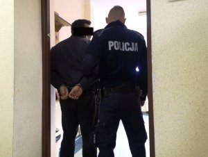 Na zdjęciu po prawej stronie stoi umundurowany policjant, po lewej mężczyzna z założonymi kajdankami na ręce trzymane z tyłu.