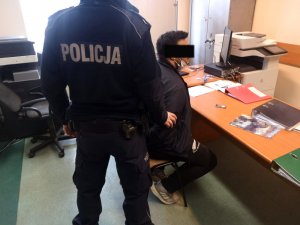 Na zdjęciu widać stojącego, odwróconego tyłem do zdjęcia, umundurowanego policjanta. Przed policjantem na krześle siedzi mężczyzna.