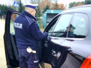 Na zdjęciu widać odwróconego tyłem do zdjęcia, umundurowanego policjanta drogówki, który stoi przy otwartych drzwiach kierowcy samochodu koloru czarnego.