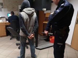 Na zdjęciu po lewej stronie stoi mężczyzna z założonymi kajdankami na ręce trzymane z tyłu, po prawej stoi umundurowany policjant. Zdjęcie jest zrobione w pomieszczeniu służbowym.