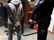 Na zdjęciu po lewej stronie stoi mężczyzna z założonymi kajdankami na ręce trzymane z tyłu, po prawej stoi umundurowany policjant.