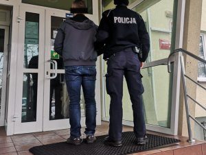 Na zdjęciu po lewej jest mężczyzna z założonymi kajdankami na ręce trzymane z tyłu, po prawej umundurowany policjant.