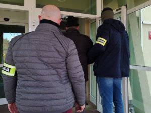 Na zdjęciu widać odwróconego tyłem do zdjęcia nieumundurowanego policjanta, który stoi na schodach. Przed nim, przed drzwiami wejściowymi do komendy, stoi drugi nieumundurowany policjant, który trzyma zatrzymanego mężczyznę.