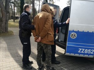 Na zdjęciu widać dwóch umundurowanych policjantów i mężczyznę z założonymi kajdankami na ręce trzymane z tyłu. Jeden z policjantów otwiera tylne drzwi do oznakowane radiowozu.