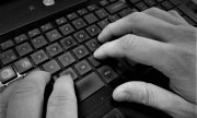 Na zdjęciu widać dwie dłonie ułożone na klawiaturze komputera. Zdjęcie jest w kolorach czarno-białych.
