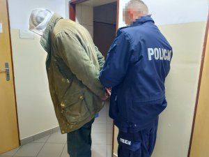 Na zdjęciu widać umundurowanego policjanta, który zakłada kajdanki mężczyźnie na ręce trzymane z tyłu.