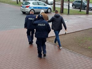 Na zdjęciu widać dwóch umundurowanych policjantów i jednego nieumundurowanego policjanta, którzy pomiędzy sobą prowadzą mężczyznę w kierunku radiowozu.