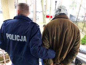 Na zdjęciu widać umundurowanego policjanta, który po prawej stronie trzyma mężczyznę z założonymi kajdankami na ręce trzymane z tyłu. Obaj są odwróceni tyłem do zdjęcia.