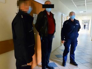 Na zdjęciu widać umundurowanego policjanta i umundurowaną policjantkę. Pomiędzy nimi stoi mężczyzna. Zdjęcie jest zrobione na korytarzu komendy.