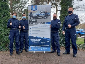 Na zdjęciu widać pięcioro, umundurowanych funkcjonariuszy, którzy stoją przy plakacie promującym zawód policjanta.