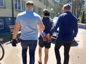 Na zdjęciu widać dwóch nieumundurowanych policjantów, którzy pomiędzy sobą prowadzą w kierunku radiowozu mężczyznę. Mężczyzna ma założone kajdanki na ręce trzymane z tyłu.