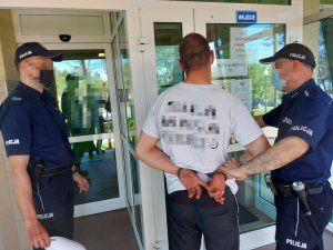 Na zdjęciu widać dwóch umundurowanych policjantów. Jeden z nich trzyma mężczyznę z założonymi na ręce trzymane z tyłu kajdankami. Zdjęcie jest zrobione przed wejściem do budynku otwockiej komendy.