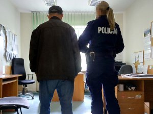 Na zdjęciu po prawej stronie stoi umundurowana policjantka, po lewej mężczyzna. Oboje są odwrócenie tyłem do zdjęcia. Zdjęcie jest wykonane w pokoju służbowym.