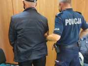 Na zdjęciu po lewej stronie stoi mężczyzna, po prawej umundurowany policjant. Obaj są odwróceni tyłem do zdjęcia.  Zdjęcie jest wykonane w pokoju służbowym.