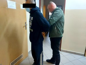 Na zdjęciu widać nieumundurowanego policjant, który zakłada mężczyźnie kajdanki na ręce trzymane z tyłu. Zdjęcie jest wykonane w pomieszczeniu służbowym.