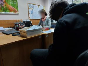 Na zdjęciu widać siedzącego na krześle mężczyznę. W tle znajduje się siedząca przy biurku nieumundurowana policjantka. Na biurku leżą dokumenty, kodeks, i laptop.