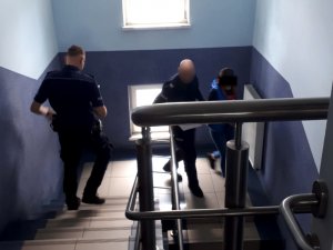 Na zdjęciu widać umundurowanych policjantów, którzy prowadzą po schodach kobietę z założonymi kajdankami na ręce trzymane z przodu.