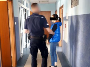 Na zdjęciu widać umundurowanego policjanta, obok niego stoi kobieta z założonymi kajdankami na ręce trzymane z przodu.