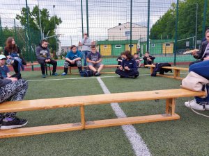 Na zdjęciu widać siedzącą po turecku na trawie umundurowaną policjantkę. Obok niej w okręgu, na ławkach siedzi młodzież.