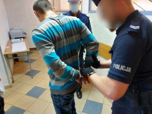Na zdjęciu widać umundurowanego policjanta, który zakłada kajdanki mężczyźnie na ręce trzymane z tyłu. Zdjęcie jest wykonane na korytarzu budynku komendy.