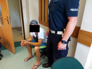 Na zdjęciu widać siedzącego na krześle mężczyznę, który ma założone kajdanki na ręce trzymane z przodu. Obok niego stoi umundurowany policjant. zdjęcie jest wykonane  w pomieszczeniu służbowym.