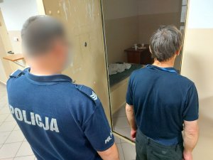 Na zdjęciu widać umundurowanego policjanta, który trzyma otwarte drzwi do policyjnej celi. Tuż przed nim stoi mężczyzna. Obydwaj są odwróceni tyłem do zdjęcia.