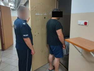Na zdjęciu widać umundurowanego policjanta, który trzyma otwarte drzwi do policyjnej celi. Tuż przed nim stoi mężczyzna.