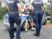 Na zdjęciu widać dwóch umundurowanych policjantów, którzy pomiędzy sobą trzymają mężczyznę z założonymi kajdankami. Osoby stoją przed radiowozem i są odwróceni tyłem do zdjęcia.