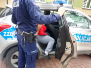 Na zdjęciu widać umundurowanego policjanta, który trzyma otwarte , tylne drzwi oznakowanego radiowozu. Na tylnej kanapie pojazdu siedzi mężczyzna.