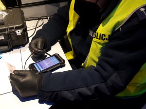 Na zdjęciu jest umundurowany policjant, który dokonuje sprawdzenia dokumentu za pomocą mobilnego terminala.