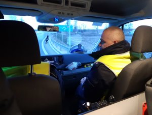 Na zdjęciu widać dwóch umundurowanych policjantów z drogówki, którzy jadąc radiowozem patrolują ulice. Zdjęcie jest zrobione wewnątrz pojazdu.