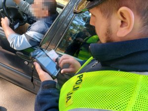 Na zdjęciu widać umundurowanego policjanta drogówki, który dokonuje sprawdzenie danych z dokumentu na terminalu mobilnym. W tle widać siedzącego za kierownicą pojazdu mężczyznę.