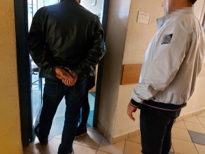 Na zdjęciu po lewej stronie stoi mężczyzna z założonymi kajdankami na ręce trzymane z tyłu, po prawej nieumundurowany policjant. Zdjęcie jest wykonane przed otwartymi drzwiami do pomieszczenia służbowego.
