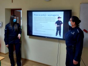Na zdjęciu widać dwie umundurowane policjantki, które omawiają slajd o treści: Praca w policji - wymagania. Zdjęcie jest wykonane w sali lekcyjnej.