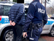 Na zdjęciu widać umundurowanego policjanta, który prowadzi mężczyznę w kierunku oznakowanego radiowozu. Mężczyzna ma założone kajdanki na ręce trzymane z tyłu.